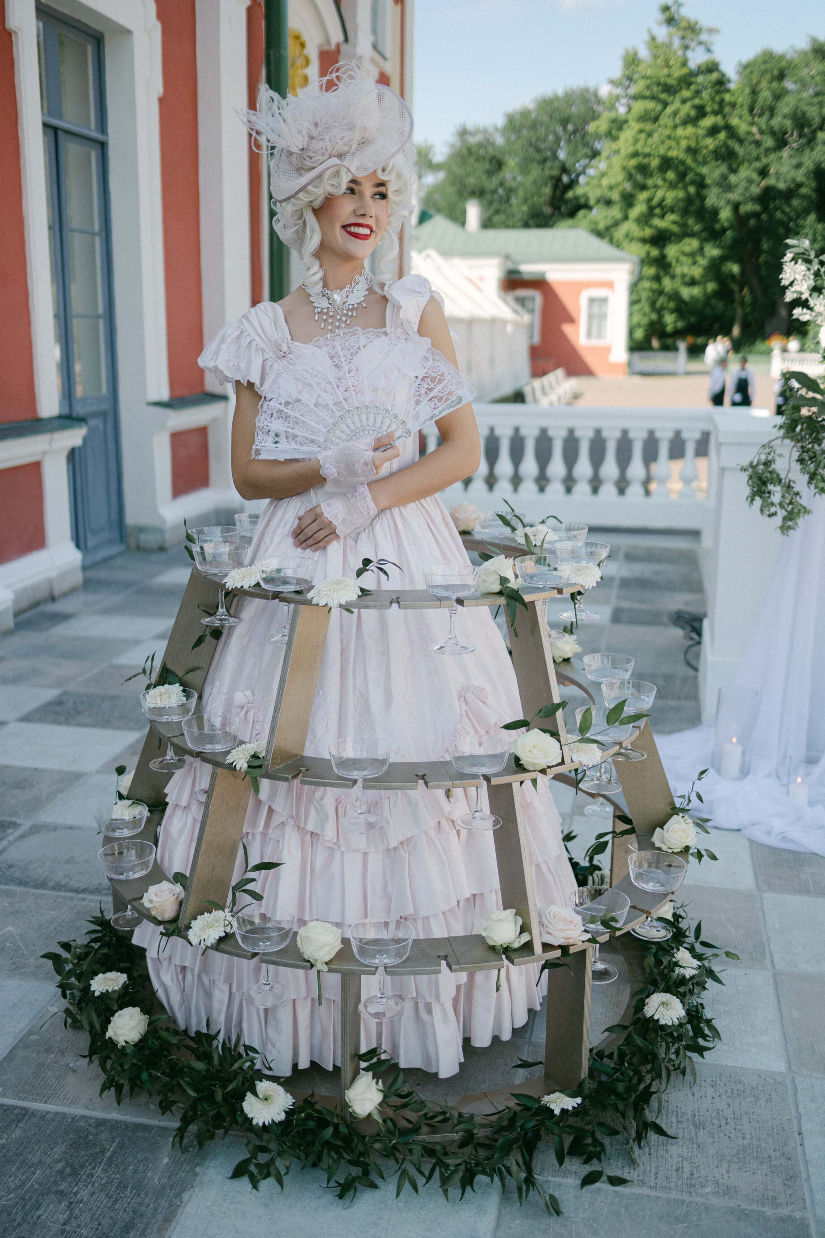 Pildil on Kadrioru lossi terrassil neiu barokk-stiilis riietuses, ümber puidust šampanjaseelik.