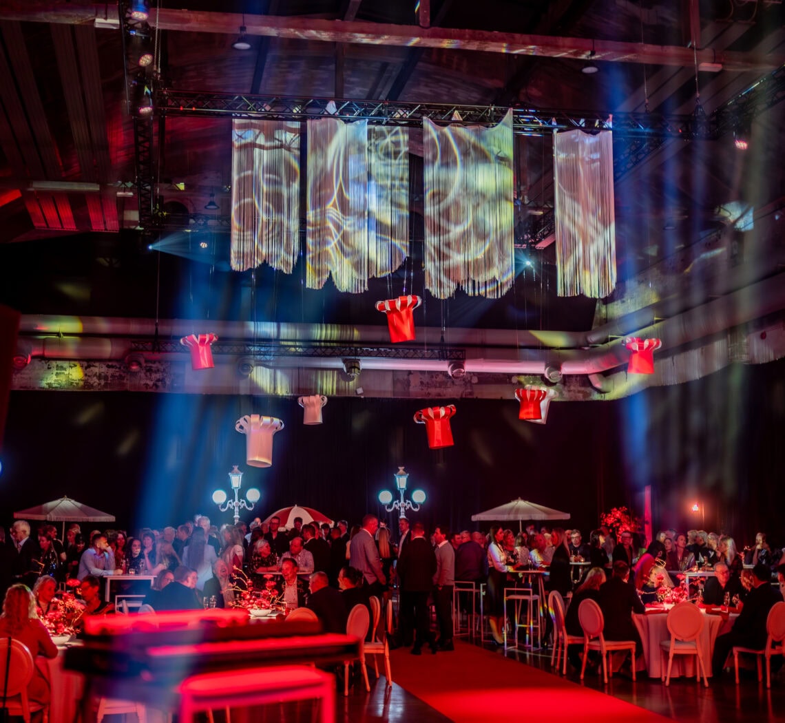 Mamma ja Saarioinen Eesti juubeliüritus Nobeli saalis, kus külalised istuvad laudade taga ning laes ripuvad suured paberist kokamütsid.